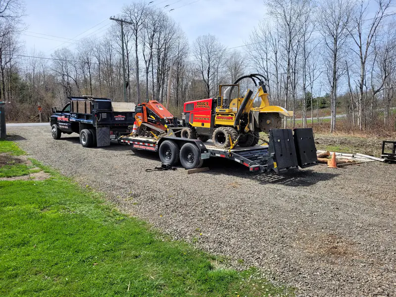 A dump truck towing a trailer carrying an orange track loader and the yellow grinder, each lettered Doug's Stump Removal