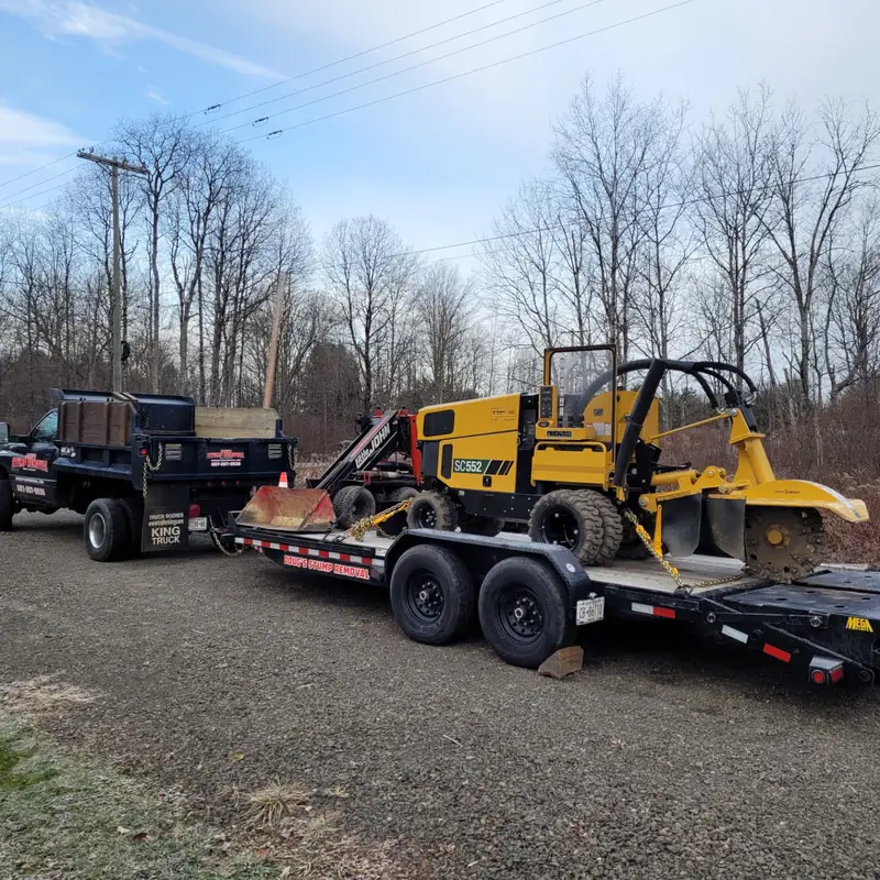 The grinder and loader on a trailer behind the black dump truck on a gravel drive, bare trees behind